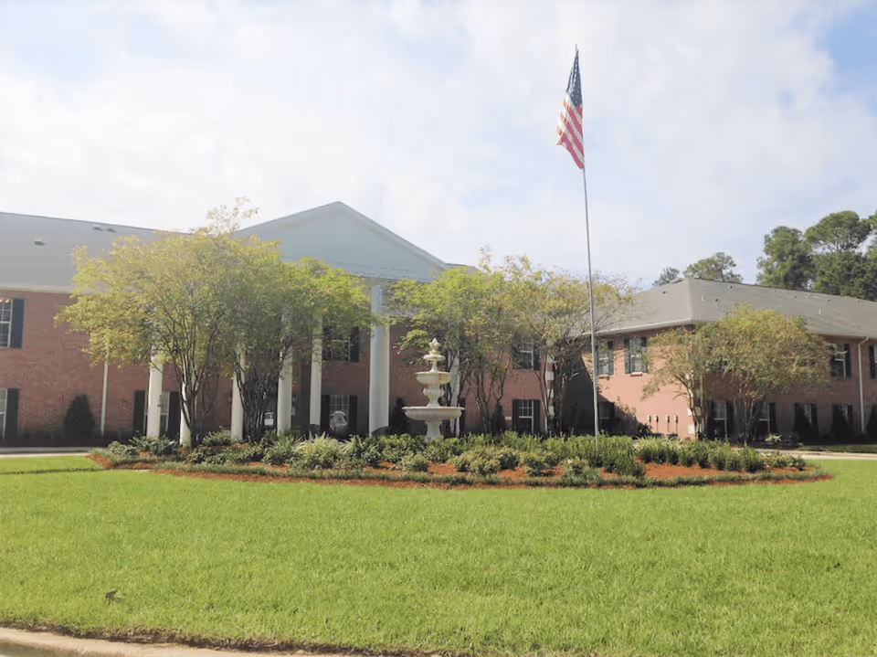Front exterior view of a two-story brick building with white columns at the entrance, surrounded by trees and a well-maintained lawn with a circular garden bed featuring a fountain and an American flag on a tall flagpole.