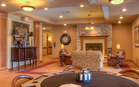 A cozy senior living common area featuring a round table with card game supplies in the foreground, a stone fireplace with a mantle and a hanging light above it, two leather armchairs, a patterned sofa, a large wall clock, and warm lighting throughout the room.