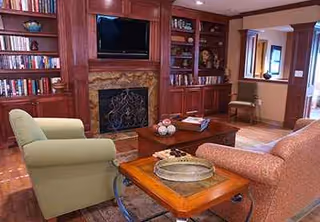 Cozy living room featuring wooden built-ins, a stone fireplace, shelves, TV, and green armchairs.