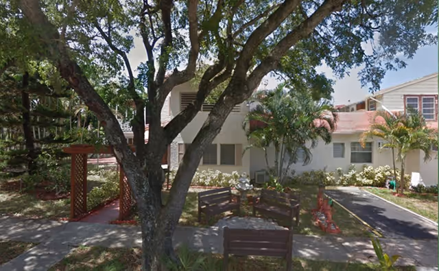 Shaded courtyard with wooden benches and a large tree in front of a low two-story residential building with palm plants.