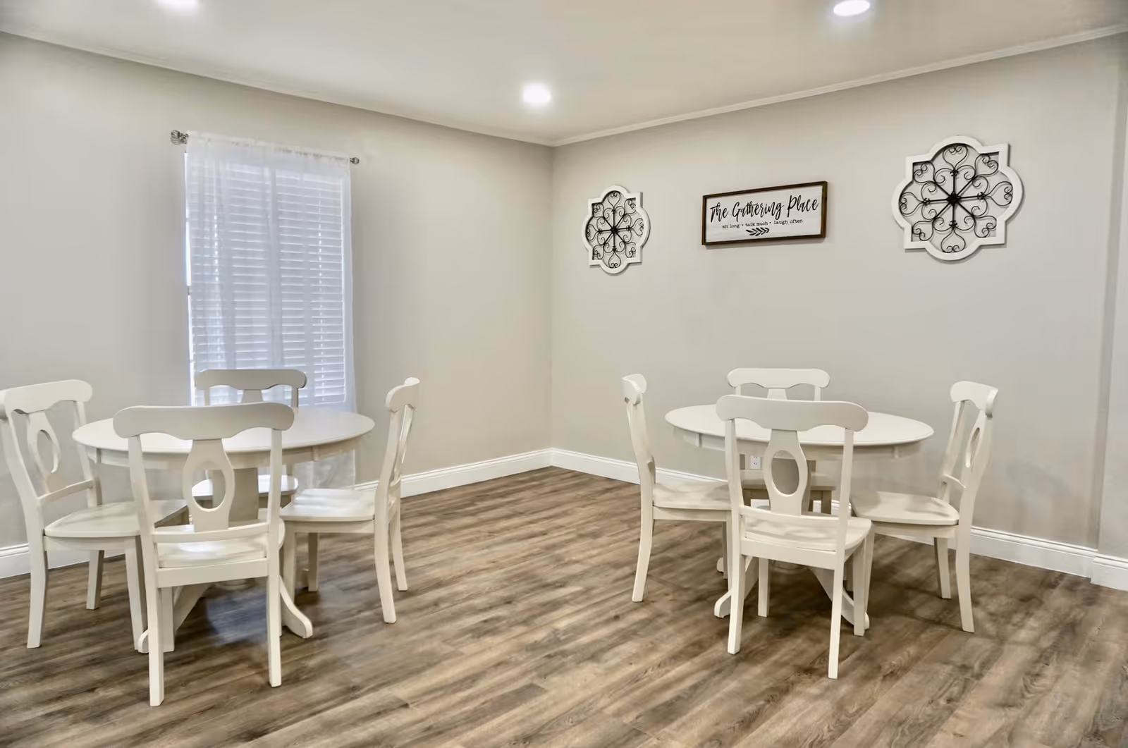 A bright room with two round white tables, each surrounded by four white wooden chairs. The floor is wood with a light brown finish. A window with white blinds and a sheer curtain is on the left wall. On the far wall, there are two decorative black wrought iron wall hangings and a sign that reads 'The Gathering Place'. The walls are painted light gray and the ceiling has recessed lighting.