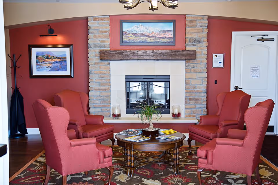 A cozy sitting area with four red upholstered armchairs arranged around a round wooden coffee table with magazines and a small plant. Behind the chairs is a stone fireplace with a wooden mantel, above which hangs a landscape painting. The walls are painted red, and there is a coat rack with a black coat on the left side. A white door is visible on the right side of the image.