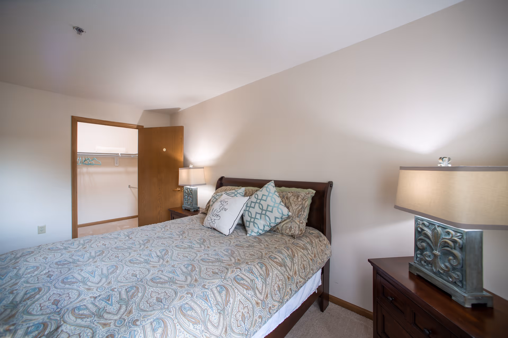 A bedroom with a neatly made bed featuring patterned bedding and three decorative pillows. There are two bedside tables with matching lamps on either side of the bed. An open door reveals a walk-in closet with hangers inside. The walls are painted light beige and the room has a calm, cozy atmosphere.