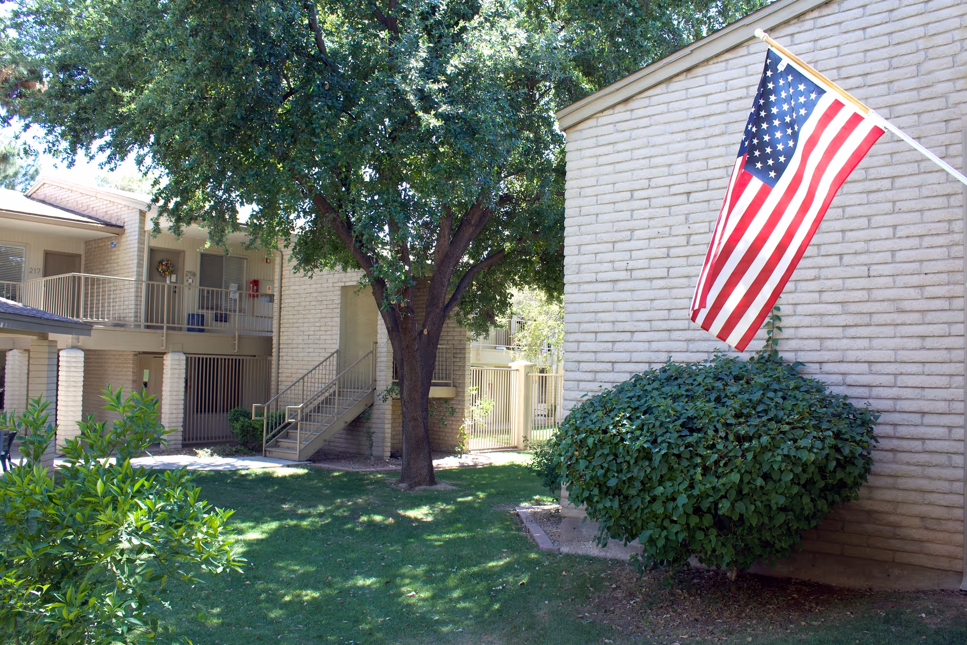 Outdoor courtyard area of a senior living facility with green grass, bushes, and a large tree. There is a beige brick building with an American flag mounted on the wall, and a staircase leading to the second floor with railings. The area is well-lit with natural sunlight.