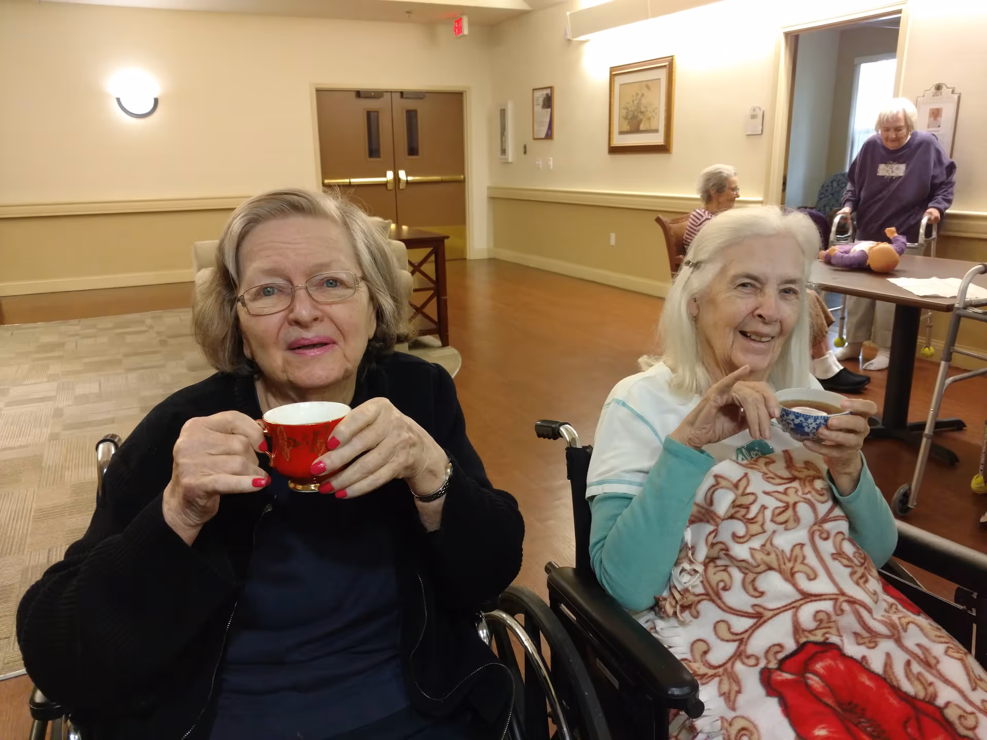 Two elderly women sitting in wheelchairs inside a senior living facility, each holding a teacup and smiling. One woman has a red teacup and glasses, the other has a blue and white teacup and a patterned blanket over her lap. In the background, two other elderly women are visible, one sitting at a table and the other using a walker.