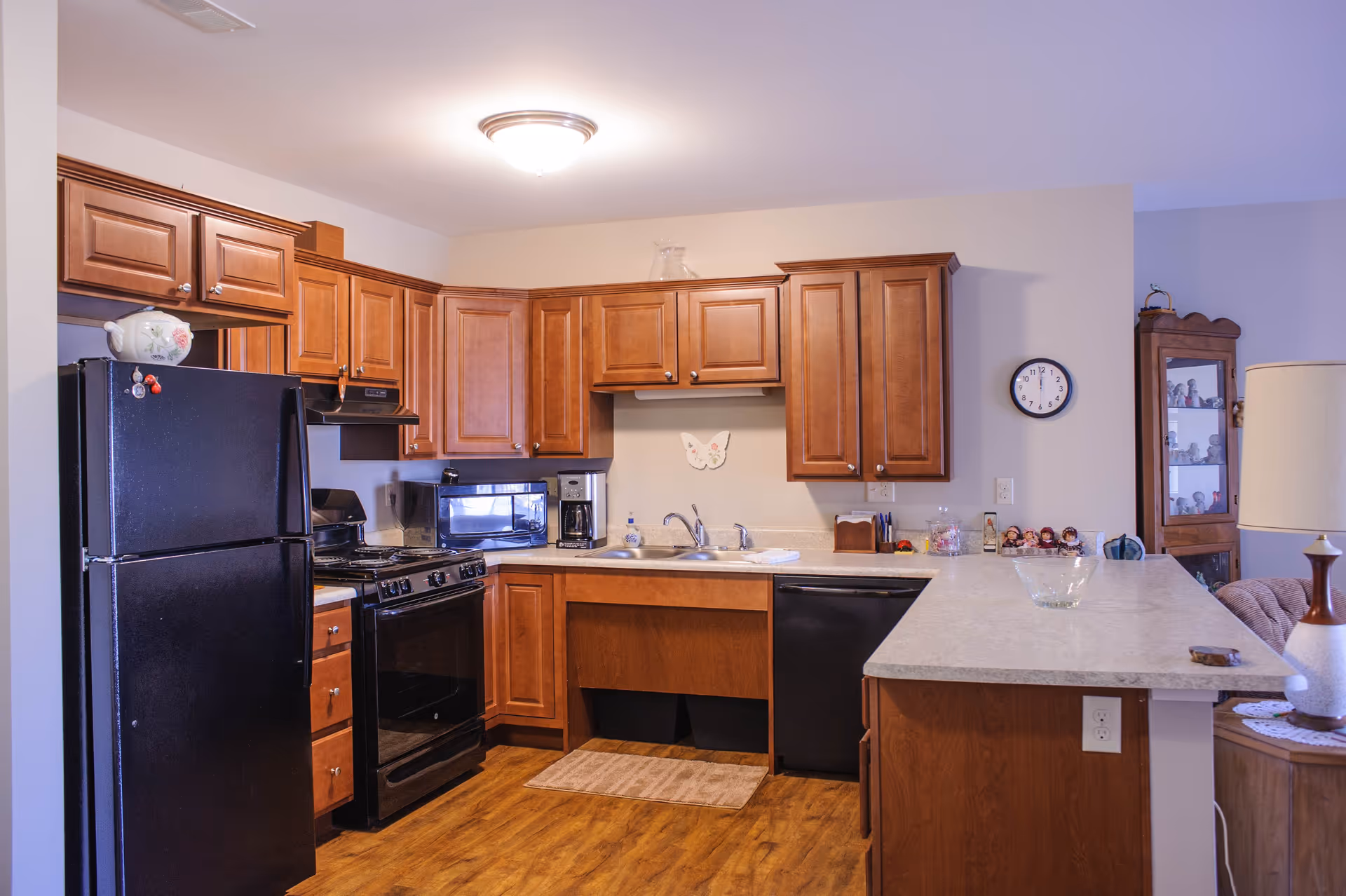 A kitchen with wooden cabinets, black refrigerator, stove, microwave, coffee maker, and dishwasher. The countertop extends into a breakfast bar with a lamp and decorative items on it. The floor is wooden, and there is a clock on the wall.