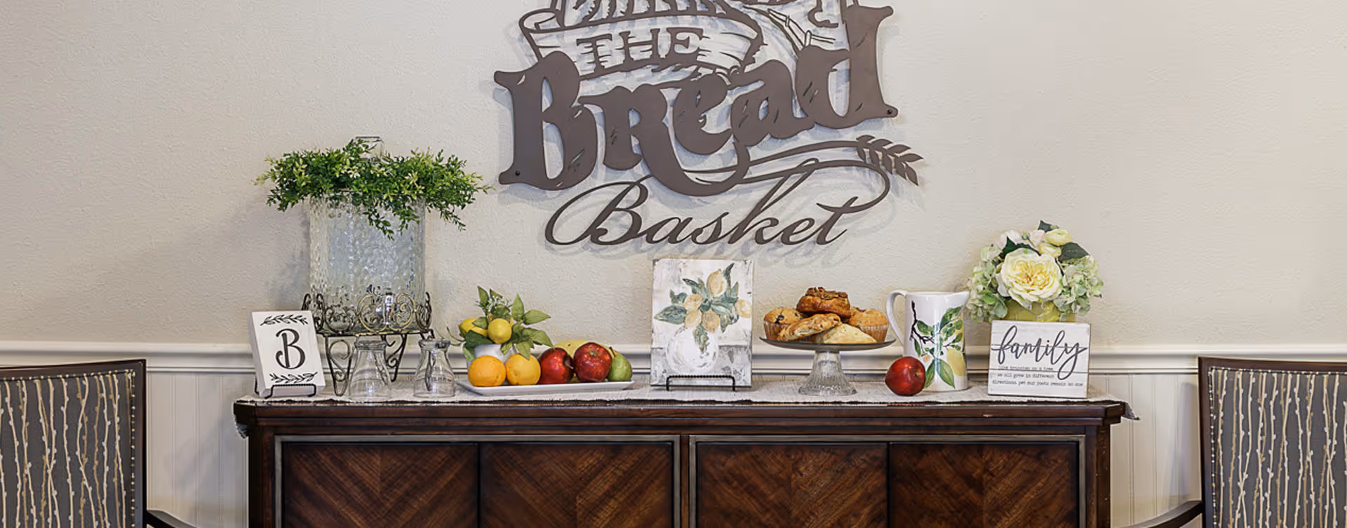 A wooden sideboard with decorative items on top including a glass water dispenser with greenery, a plate of assorted fruits, a plate with pastries, a floral pitcher, and a small flower arrangement. Above the sideboard is a wall decoration that reads 'The Bread Basket'. Two chairs with patterned upholstery flank the sideboard.