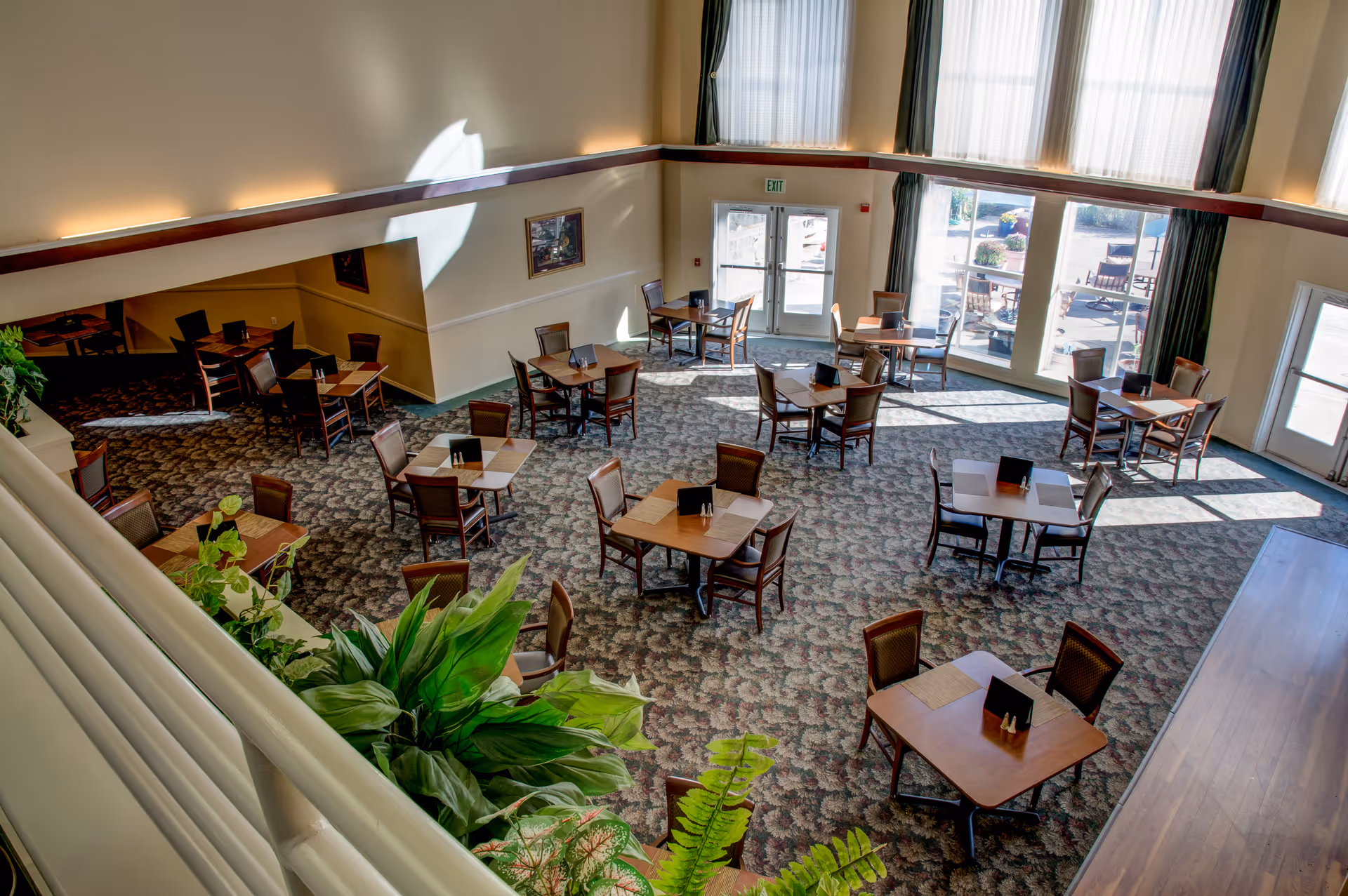 A spacious dining room with multiple square wooden tables, each surrounded by four chairs. The room has large windows letting in natural light, patterned carpet flooring, and some green plants near the railing in the foreground. There is an exit door at the far end and a painting on the wall.