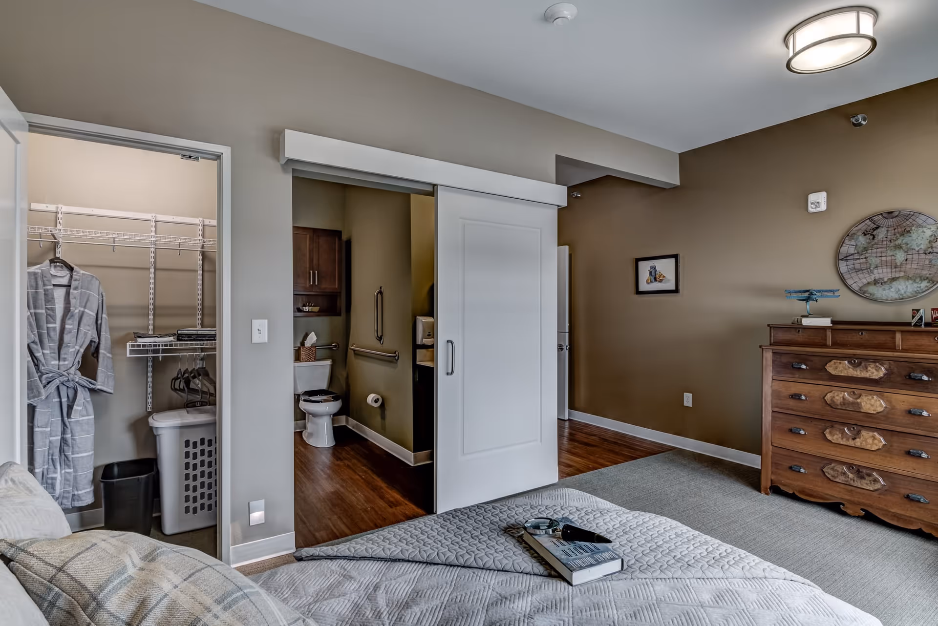 A bedroom with a bed in the foreground covered with a gray quilt and a book on top. To the left, there is an open closet with a gray robe hanging and a laundry basket. A sliding door is partially open, revealing a bathroom with a toilet, grab bars, and a wooden cabinet. On the right side of the room, there is a wooden dresser with decorative items and a wall with framed pictures and a world map decoration.
