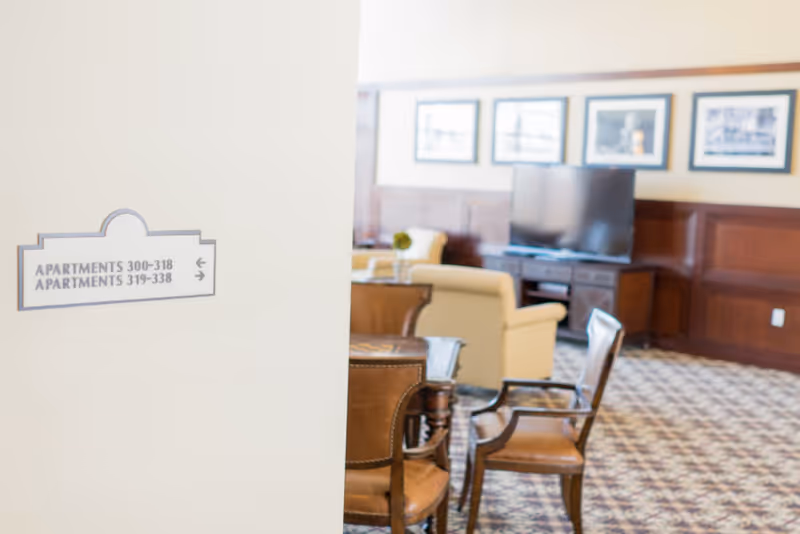 A senior living lounge with chairs, a TV, framed pictures and wood paneling visible beyond a wall sign pointing to apartment numbers.