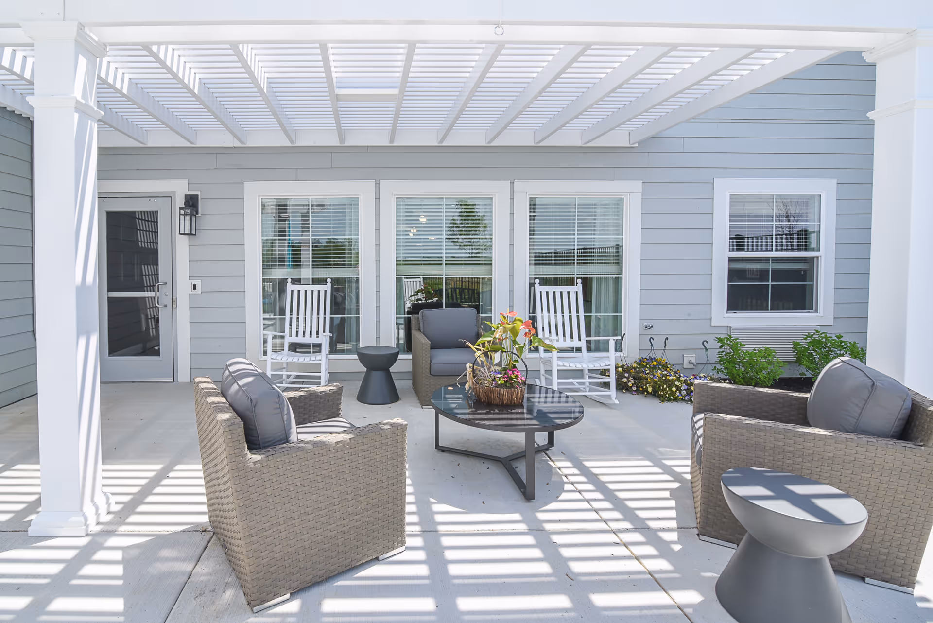 Outdoor patio area with a white pergola overhead casting shadows on the concrete floor. The space includes two wicker armchairs with gray cushions, two white rocking chairs, a small black side table, and a black coffee table with a flower arrangement. The background shows a gray building wall with windows and a door.