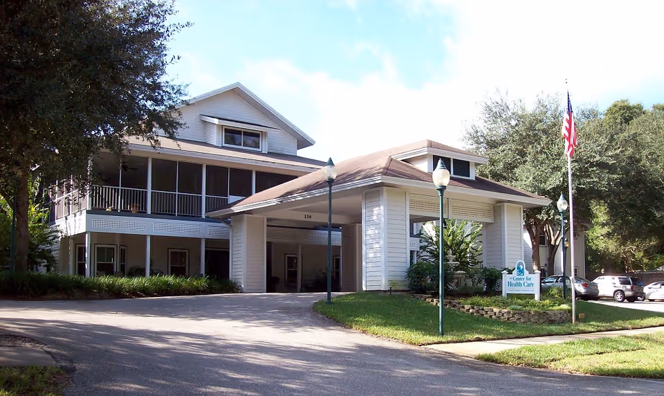 Exterior view of a two-story senior living facility building with a covered entrance, surrounded by trees and a parking area. There is a sign near the entrance that reads 'Center for Health Care' and an American flag on a flagpole.