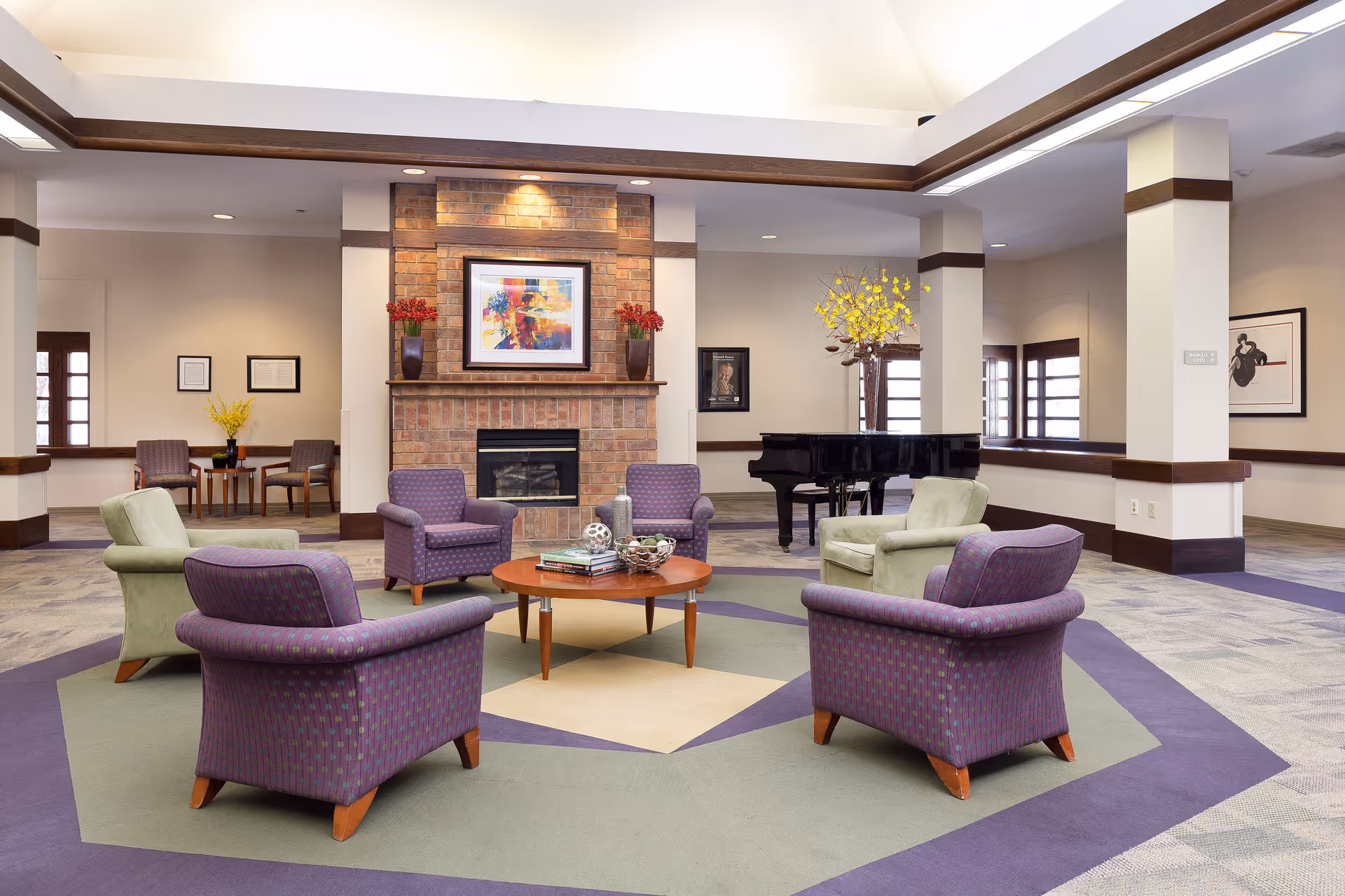 A spacious senior living lounge with armchairs arranged around a central coffee table, a brick fireplace, and a grand piano.