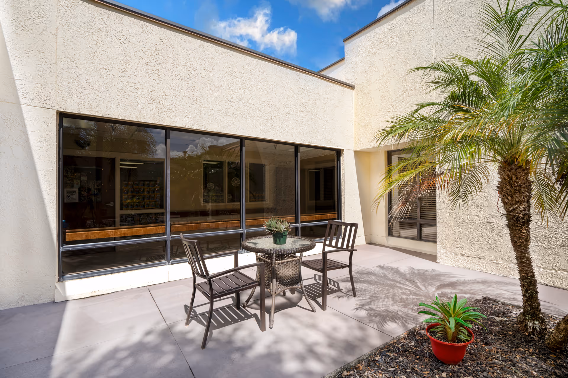 Outdoor patio area at Heritage Park Health Center with a round glass-top table and three chairs. There is a small potted plant on the table and a palm tree with another potted plant in a red container nearby. The patio is surrounded by beige stucco walls and large windows reflecting the sky and surroundings.