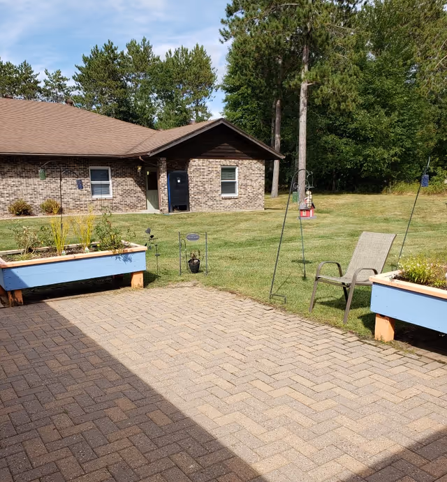 Outdoor patio area with brick pavers, two raised garden beds painted blue, a single chair, bird feeders, and a grassy lawn with trees and a brick building in the background under a partly cloudy sky.