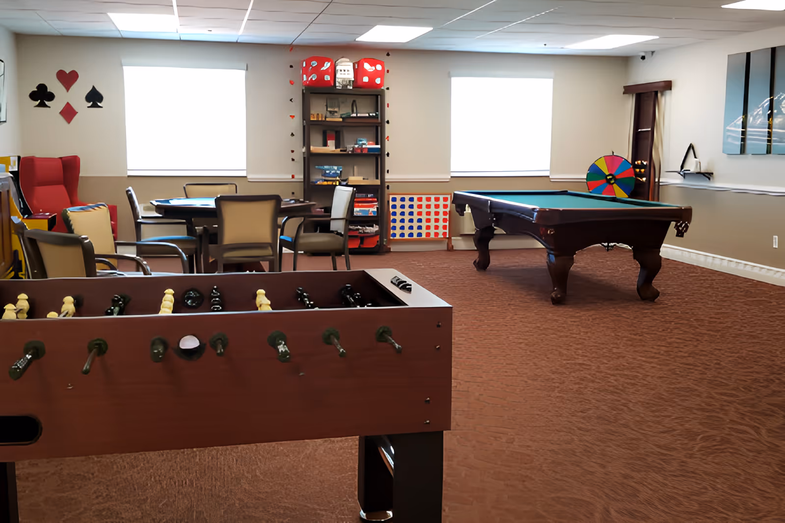 A recreational room with a foosball table in the foreground, a pool table in the middle, and a round table with chairs near two windows. The room has a bookshelf with board games and a colorful spinning wheel against the wall. The walls are decorated with card suit symbols and the carpet is brown.