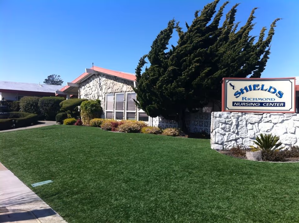 Front lawn and entrance of the Shields-Richmond Nursing Center showing a stone building, large windblown tree, and the facility sign on the right.