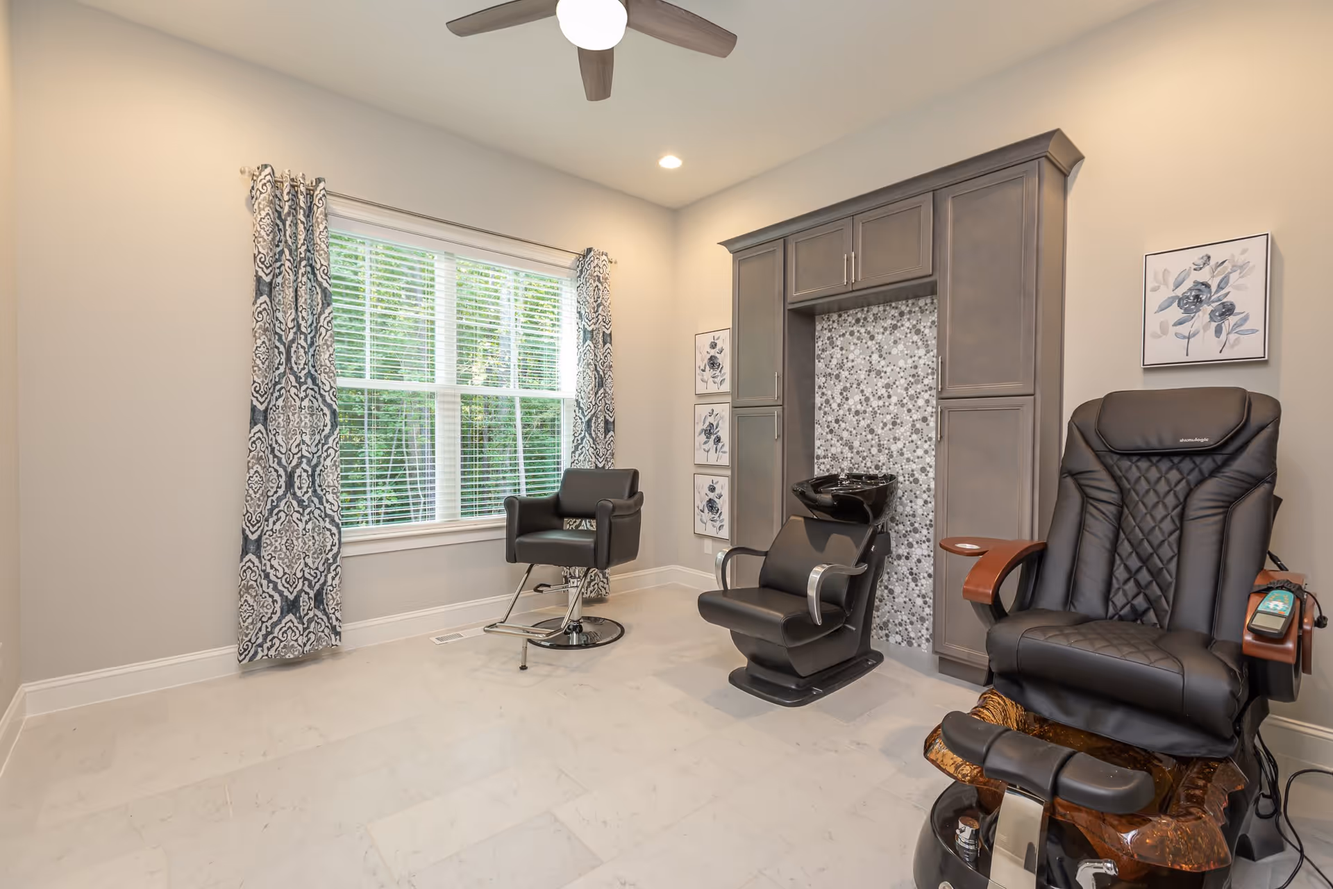 A bright and clean salon room with a large window covered by patterned curtains, a black salon chair with a hair washing basin, a black pedicure chair with wooden armrests, and a ceiling fan above. The walls are light-colored with floral artwork and built-in cabinetry surrounding the hair washing area.