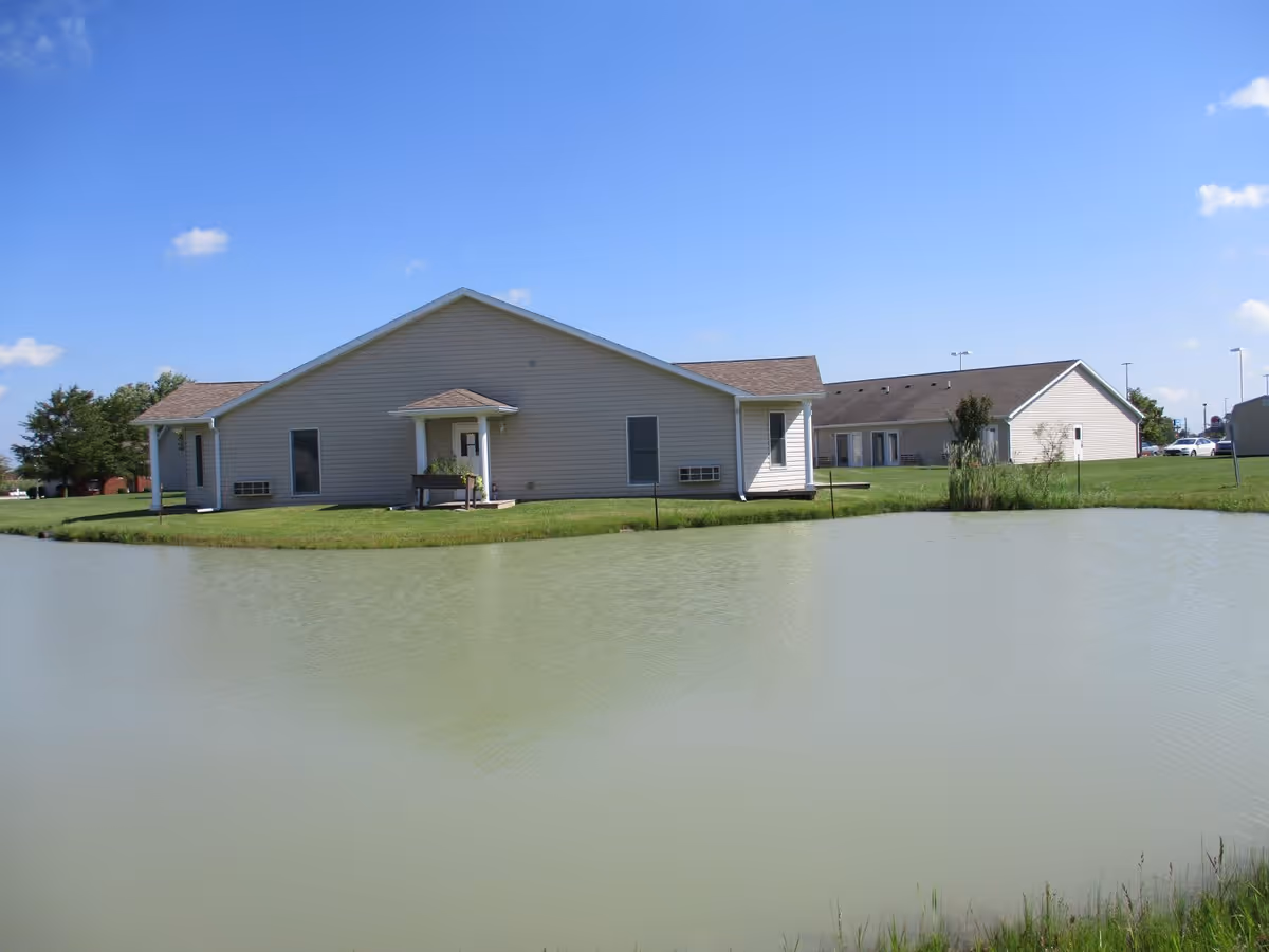 View of a single-story building with beige siding and a small covered porch, situated next to a pond with greenish water under a clear blue sky.