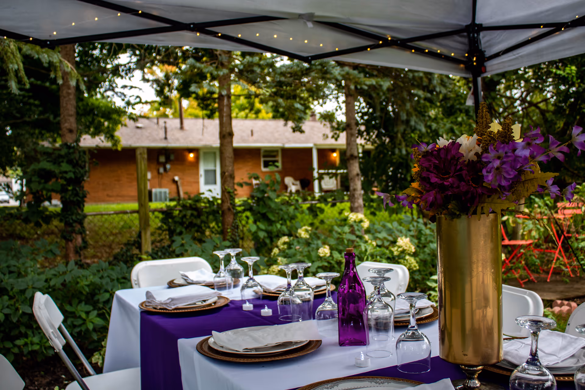 Outdoor table set for a meal under a canopy with purple accents, glassware, and a tall gold vase of flowers in a backyard.