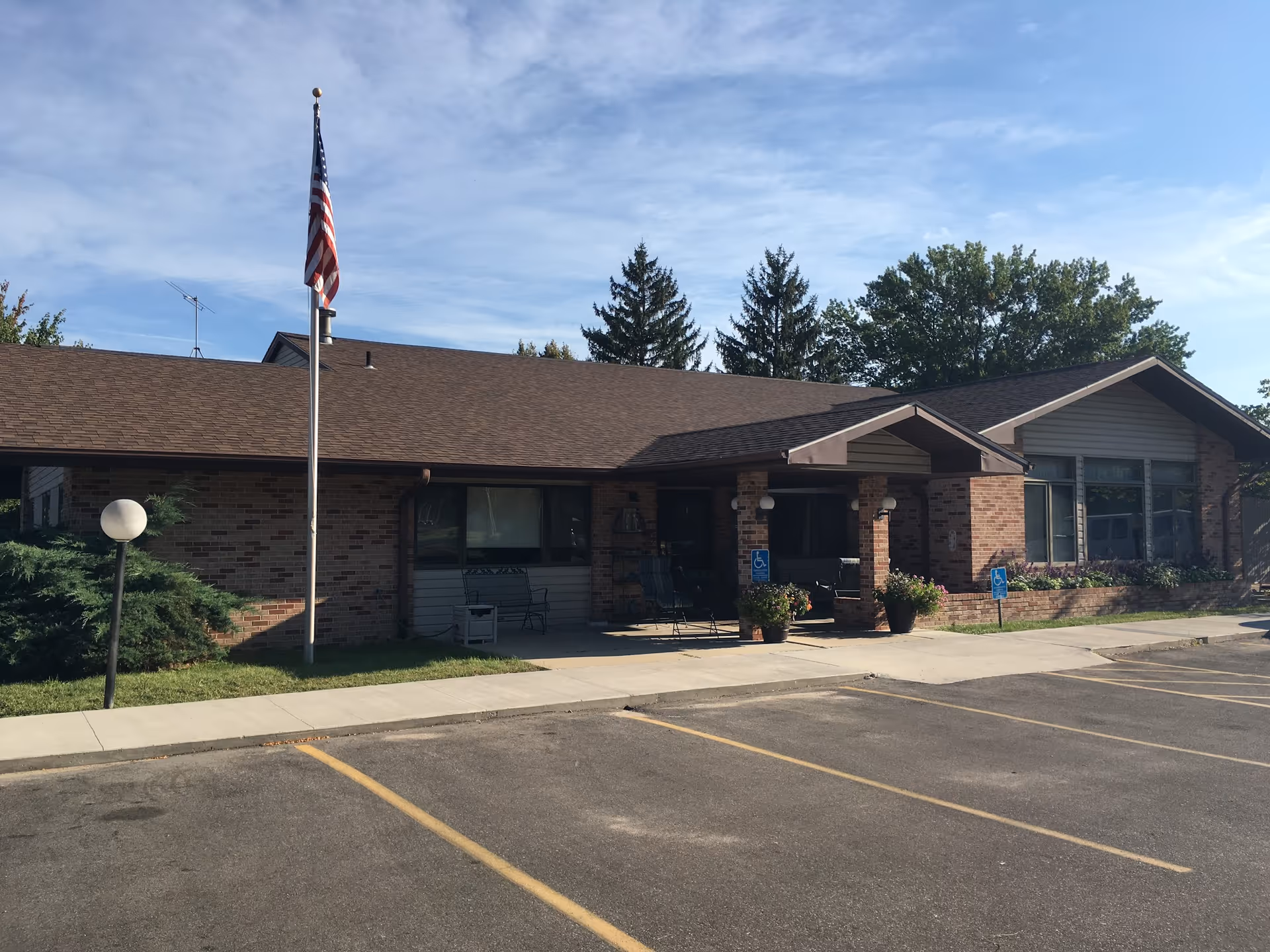 Exterior view of a single-story brick building with a brown roof, an American flag on a flagpole, and a parking lot with marked spaces in front. There are some shrubs and potted plants near the entrance, which has a covered porch area with seating. The sky is partly cloudy.