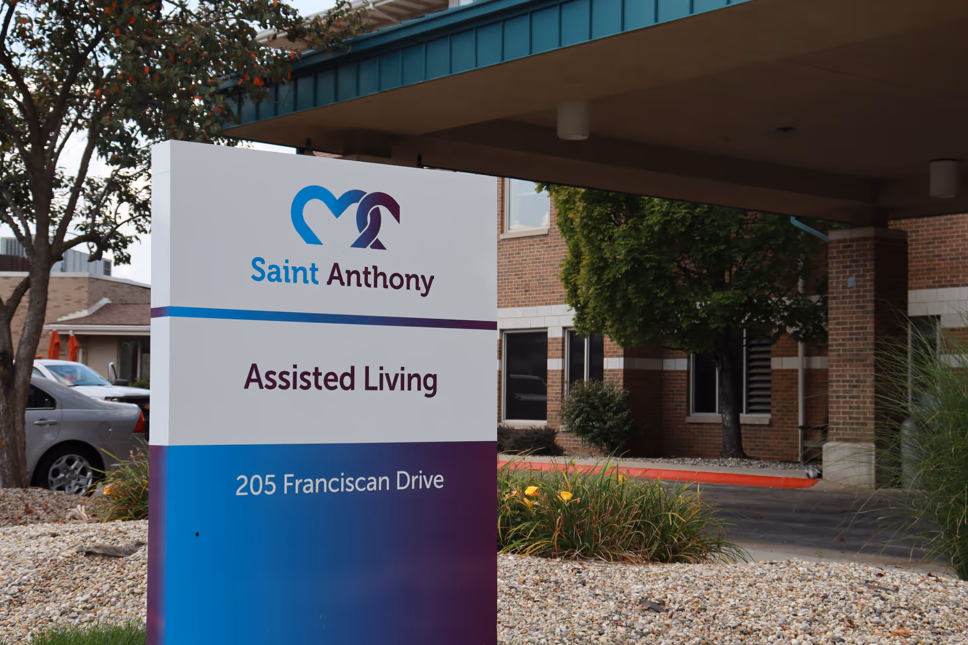 Outdoor view of the entrance area of Saint Anthony Assisted Living facility with a sign displaying the name, type of care, and address 205 Franciscan Drive. The building exterior is visible with brick walls, windows, and some greenery including trees and plants.
