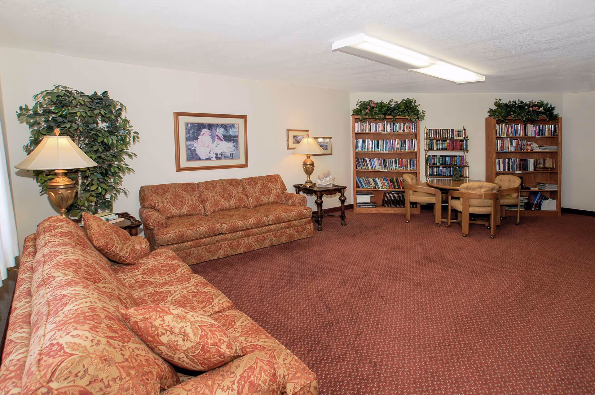 Spacious senior living lounge with patterned sofas, lamps, bookshelves and a small table with chairs.