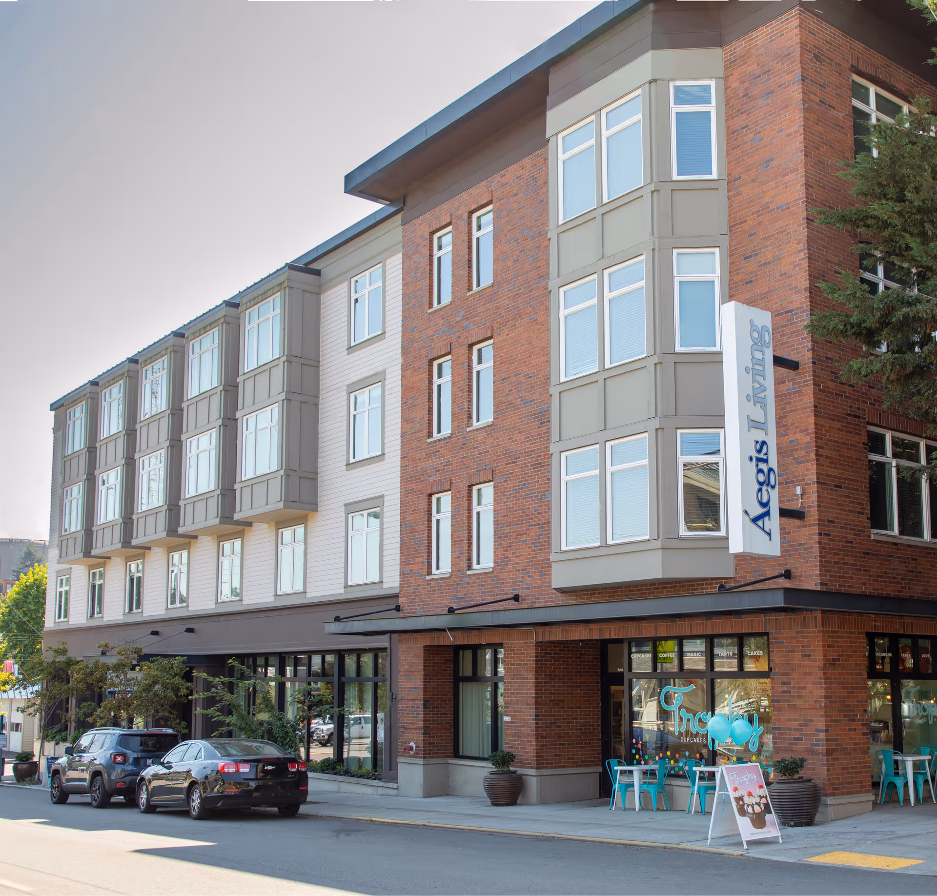 Exterior view of a multi-story brick and siding building with large windows and a sign that reads 'Aegis Living'. There are two cars parked on the street in front of the building and a small outdoor seating area with turquoise chairs and balloons near the entrance.