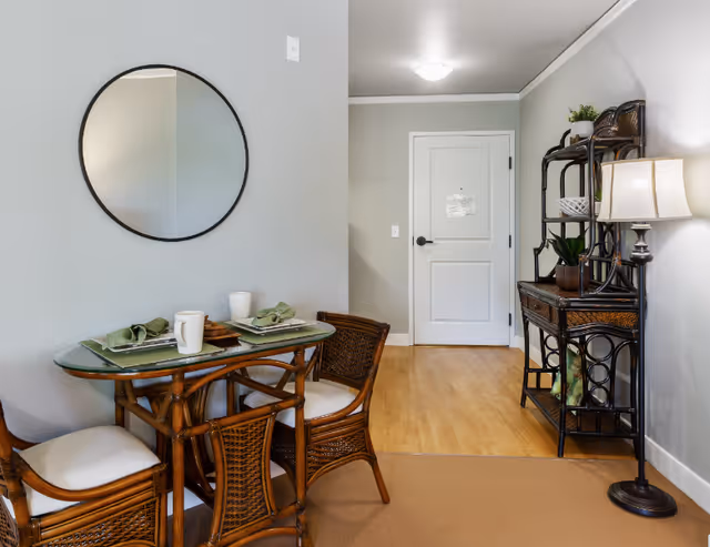 Small dining nook with a glass-top table and wicker chairs, a round wall mirror, shelving unit, and an entry door in a softly lit interior.