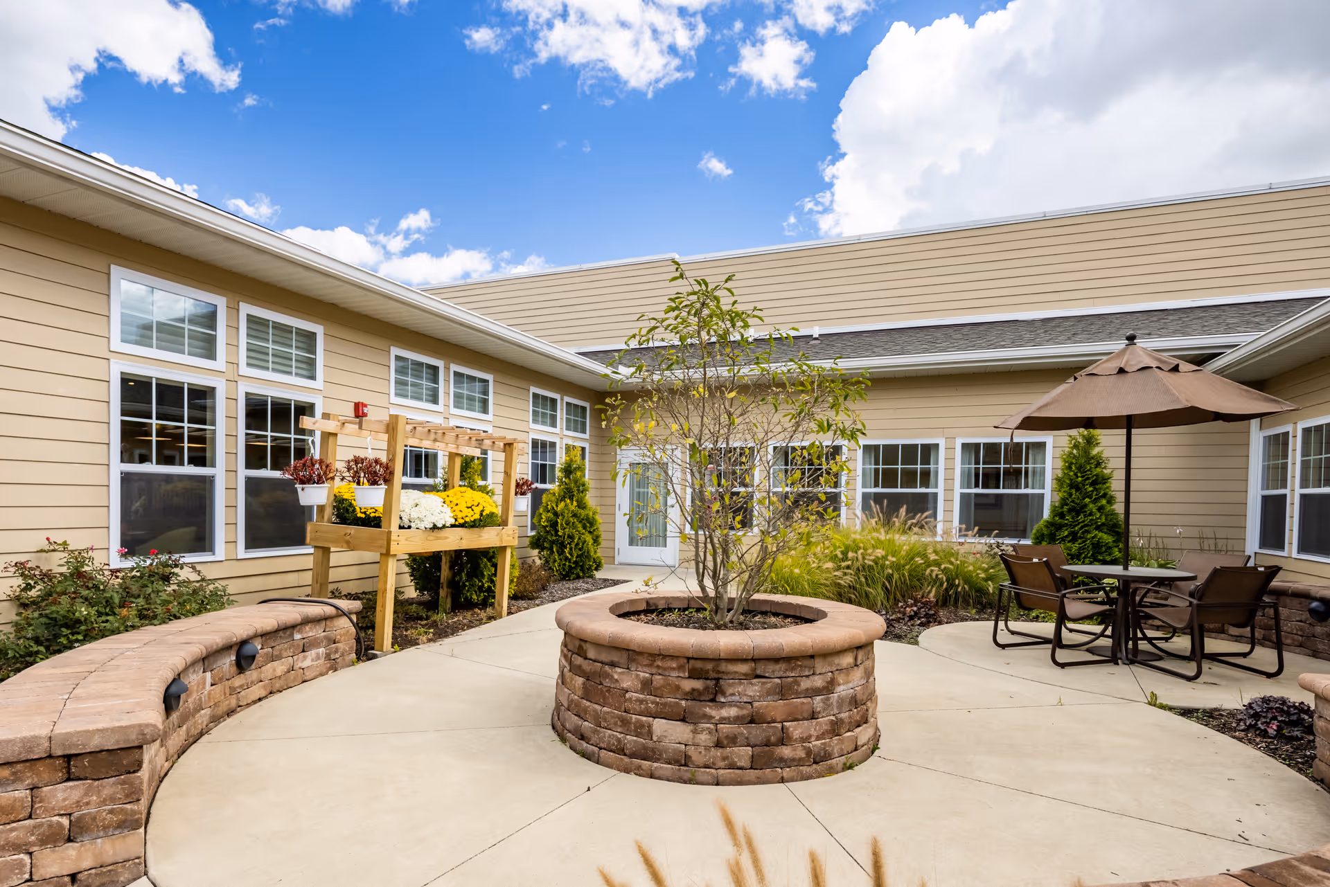 Sunny courtyard at a memory care facility featuring a central circular brick planter, curved brick seating, a patio table with umbrella, and raised flower beds against a beige building.