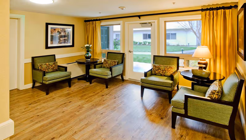 A cozy sitting area with four green upholstered armchairs arranged around two small round black tables. One table has a vase with flowers and the other has a lamp and decorative bowl. The room has wood flooring, yellow walls, a large window with yellow curtains, and a glass door leading outside to a grassy area.