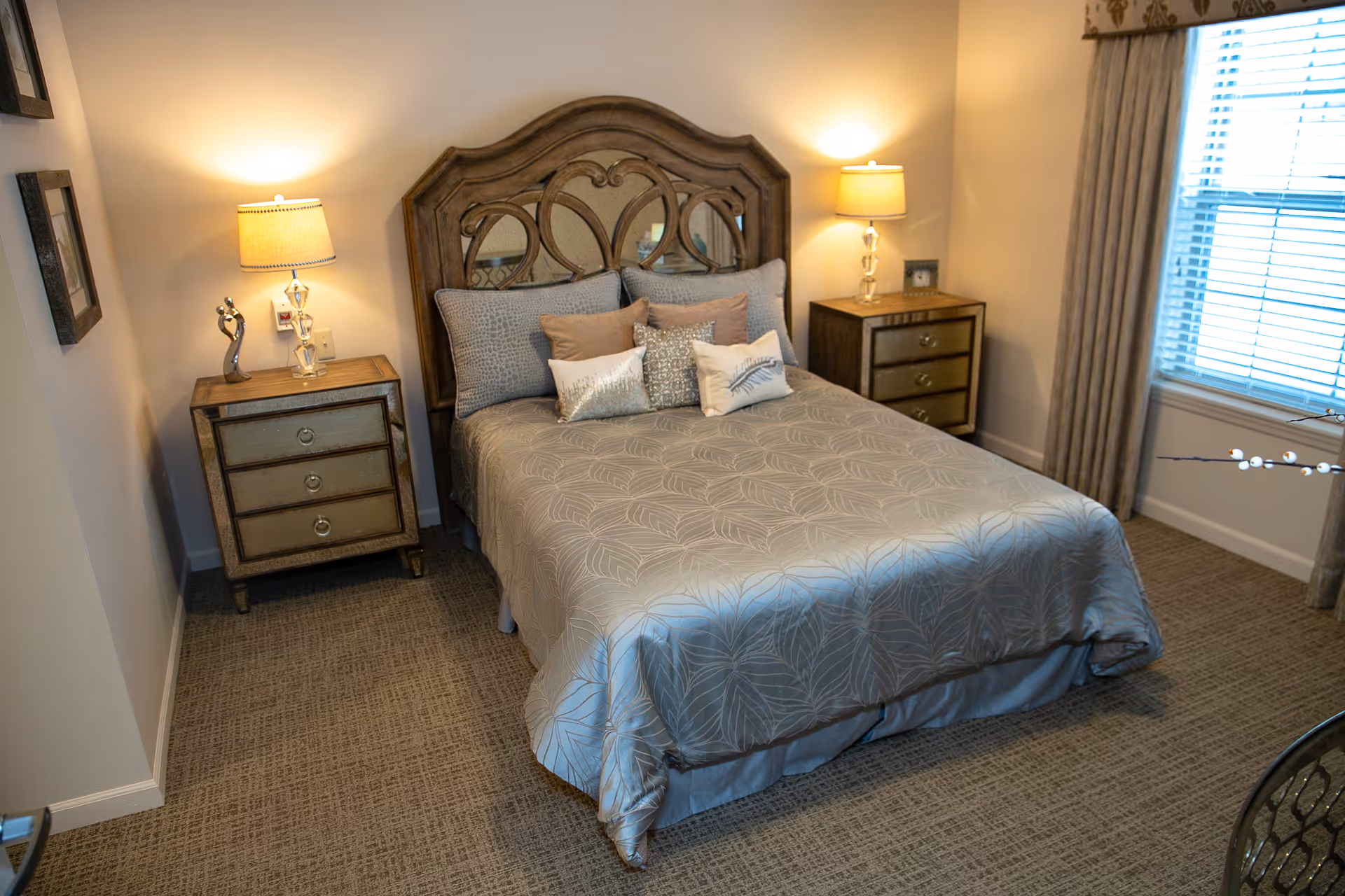 A neatly made bed with a decorative wooden headboard and multiple pillows in a softly lit bedroom. There are two matching bedside tables with lamps on each side of the bed. A window with closed blinds and curtains is visible on the right side of the room. The floor is carpeted and the walls are painted a light color.