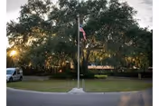 An American flag on a flagpole centered in a circular driveway with large oak trees and a low building partially visible behind, and a van parked at left.