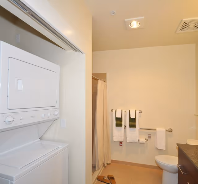 A bathroom with a stacked washer and dryer unit on the left, a shower with a beige curtain, a toilet, and a countertop with a sink. There are three towels hanging on the wall, two white with green stripes and one plain white. A pair of slippers is on the floor near the shower.