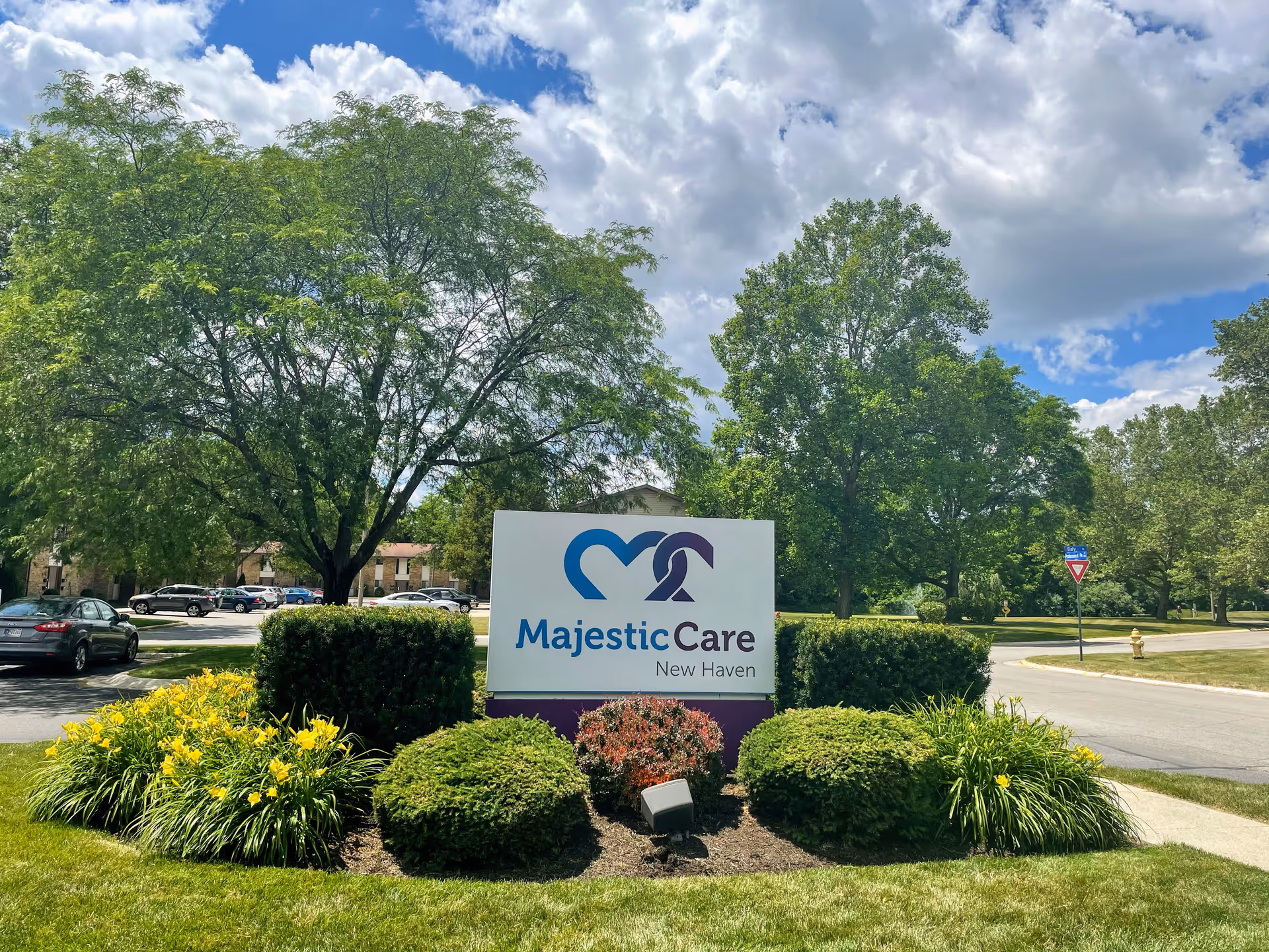 Outdoor view of a landscaped area with green bushes and yellow flowers surrounding a sign that reads 'Majestic Care New Haven' under a partly cloudy sky with trees and a road in the background.