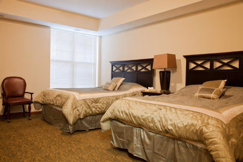 A senior living facility bedroom with two twin beds featuring dark wooden headboards and beige bedding. There is a bedside table between the beds with a lamp and a tissue box. A single wooden chair with a cushioned seat is placed near a window with closed blinds.