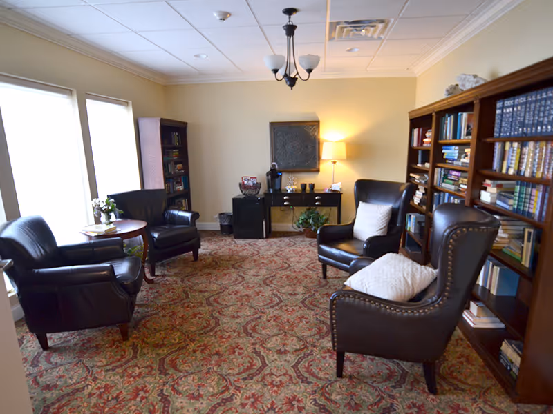 Cozy communal lounge with leather armchairs, bookshelves, a patterned carpet, and a lamp-lit side table.