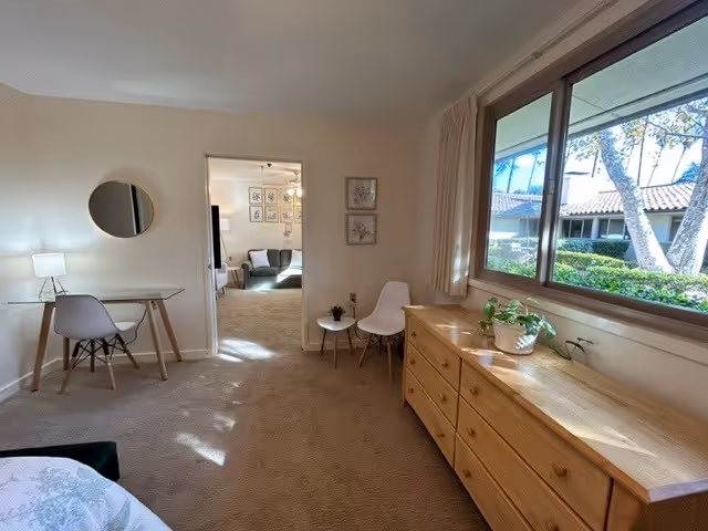 A bright bedroom in a senior living facility with a large window showing outdoor greenery. The room features a wooden dresser with plants on top, a small white chair next to a small round table, and a glass desk with a lamp and chair. Through an open doorway, a living room area with a couch and framed artwork on the wall is visible.