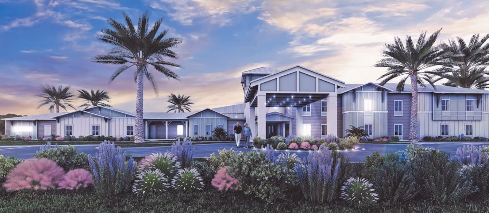 Exterior front view of a senior living building with a covered entrance, palm trees, and landscaped gardens at sunset.