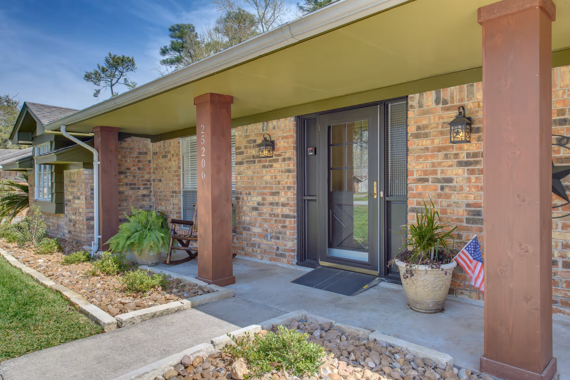 Front porch of a brick building with a black door, two wooden pillars, a rocking chair, potted plants, and an American flag. The porch has a concrete floor and is surrounded by a landscaped garden with rocks and greenery.