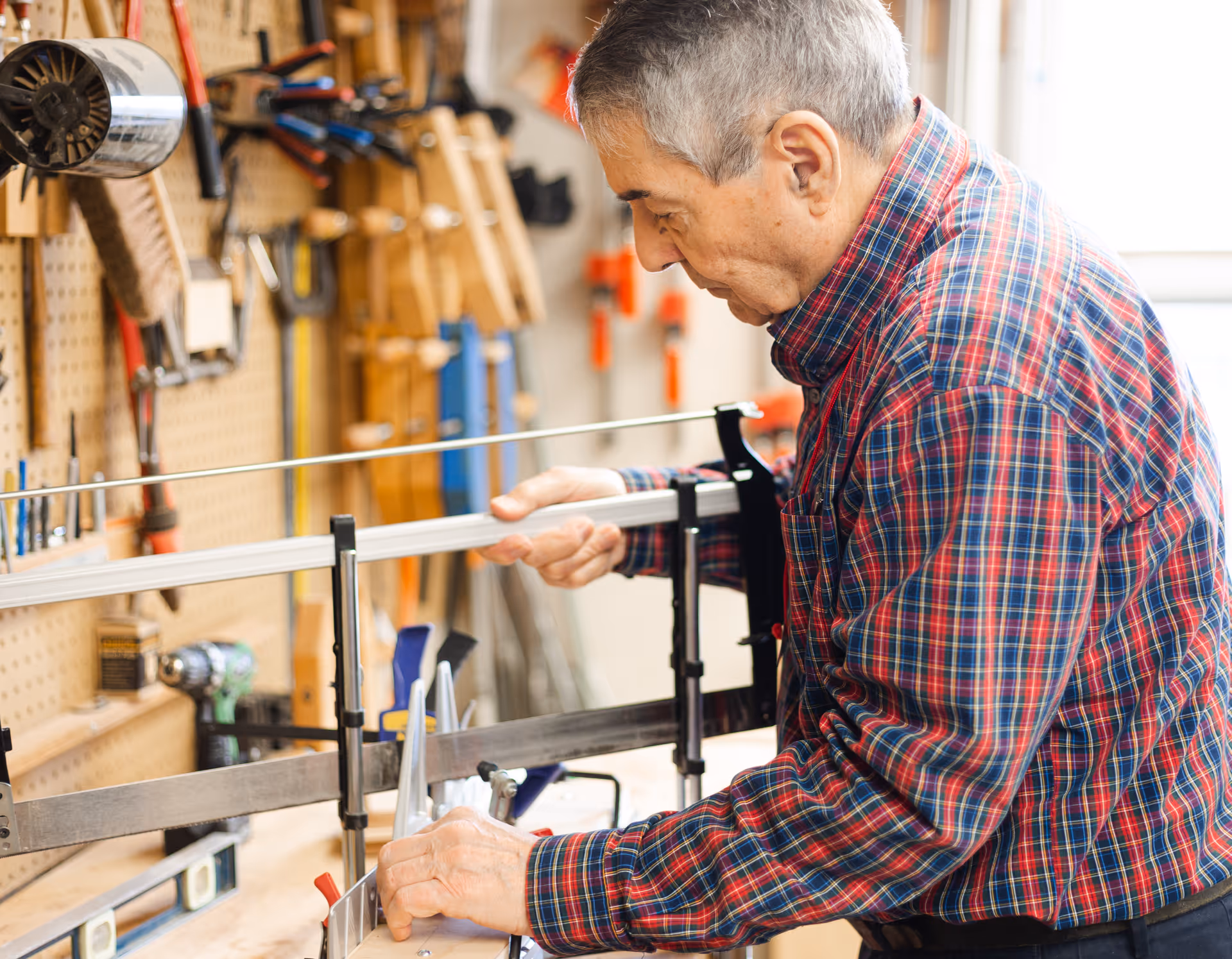 An elderly man wearing a red and blue plaid shirt is working with a woodworking tool in a workshop. Various tools and equipment are hanging on a pegboard wall in the background.
