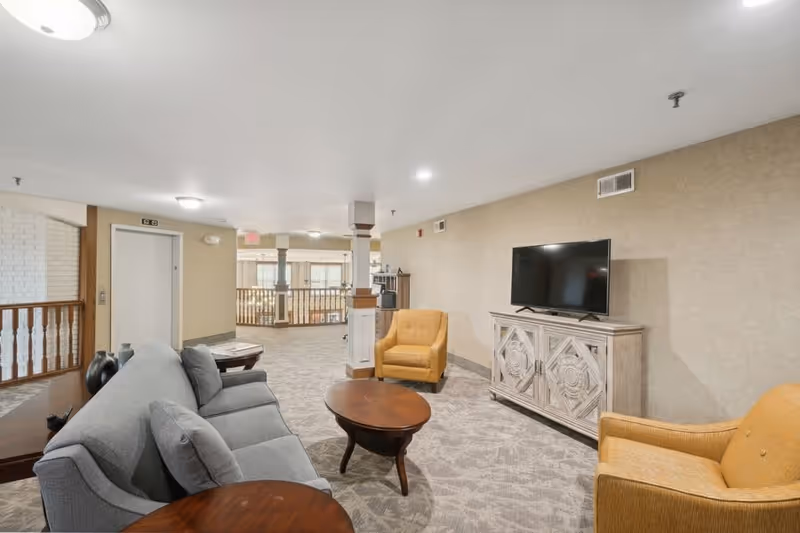 A spacious living room area in a senior living facility with a gray sofa, two mustard yellow armchairs, a wooden coffee table, and a TV on a decorative wooden cabinet. The room has beige walls, carpeted floors, and ceiling lights. There is a railing and an elevator door visible in the background.