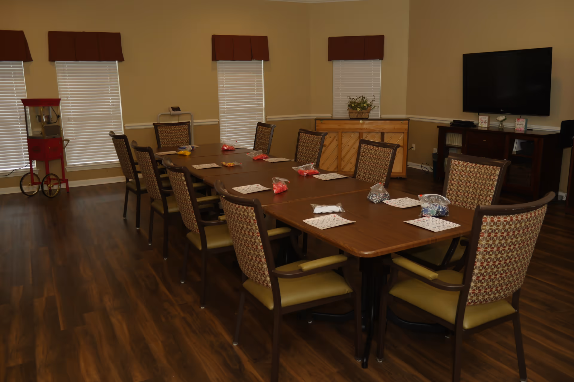 A well-lit room with a long rectangular table surrounded by ten cushioned chairs. On the table are bingo cards and small bags of colorful bingo chips. The room has wooden flooring, four windows with closed blinds and valances, a red popcorn machine in the corner, a piano with a flower pot on top, and a flat-screen TV mounted on the wall above a cabinet.