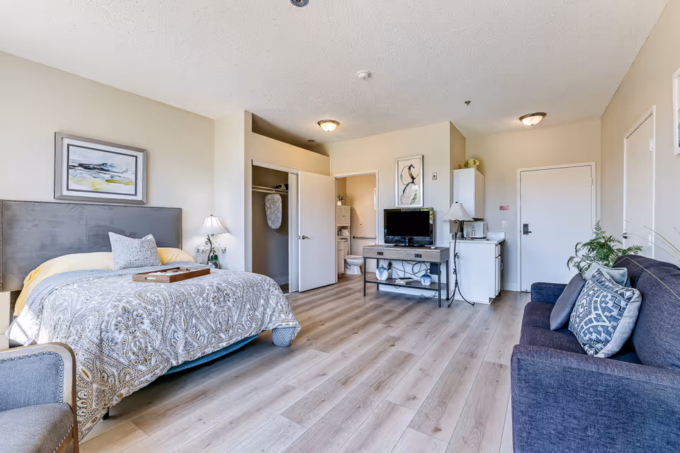 A furnished senior living bedroom with a bed, seating area, closet, and TV.