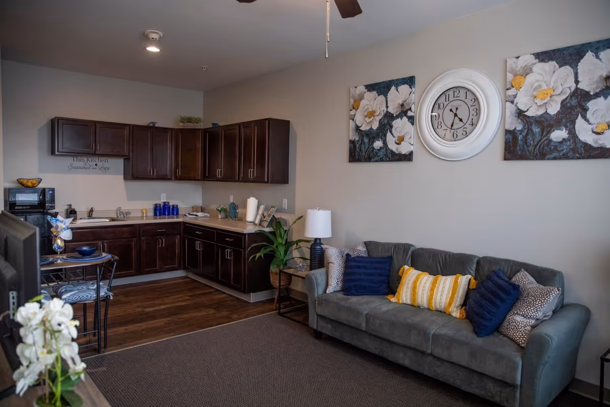 Open living area with a gray couch and decorative pillows under a wall clock, adjacent to a small kitchenette with dark cabinets.