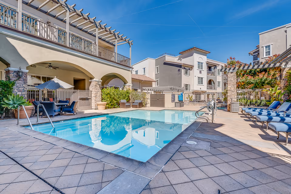 Outdoor swimming pool and surrounding patio with lounge chairs, seating areas, and adjacent multi-story residential building.