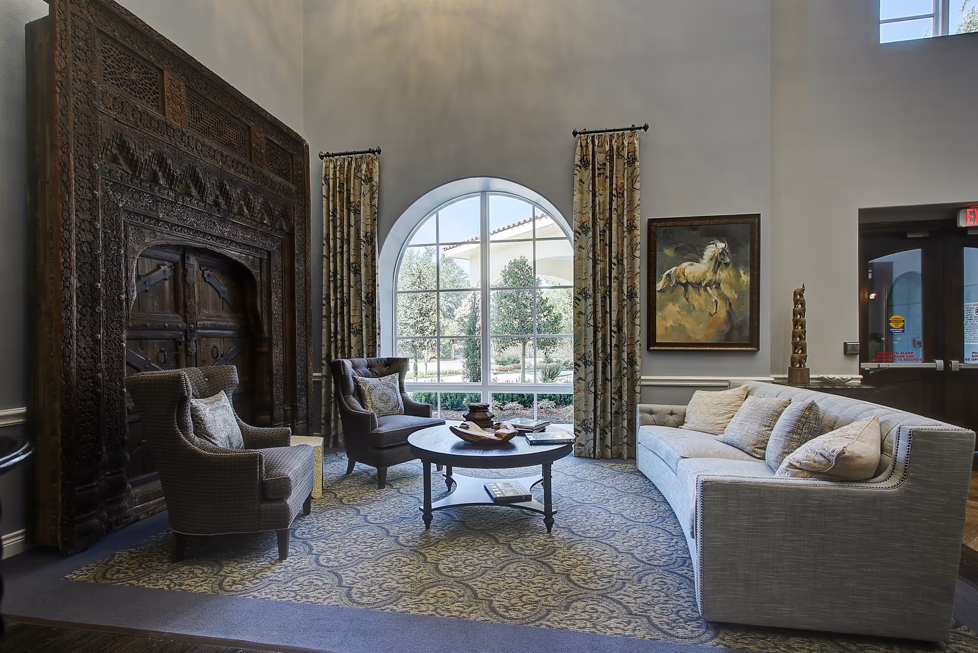 Comfortable seating area with a sofa, two armchairs, a round coffee table, ornate carved wooden paneling, and a large arched window.