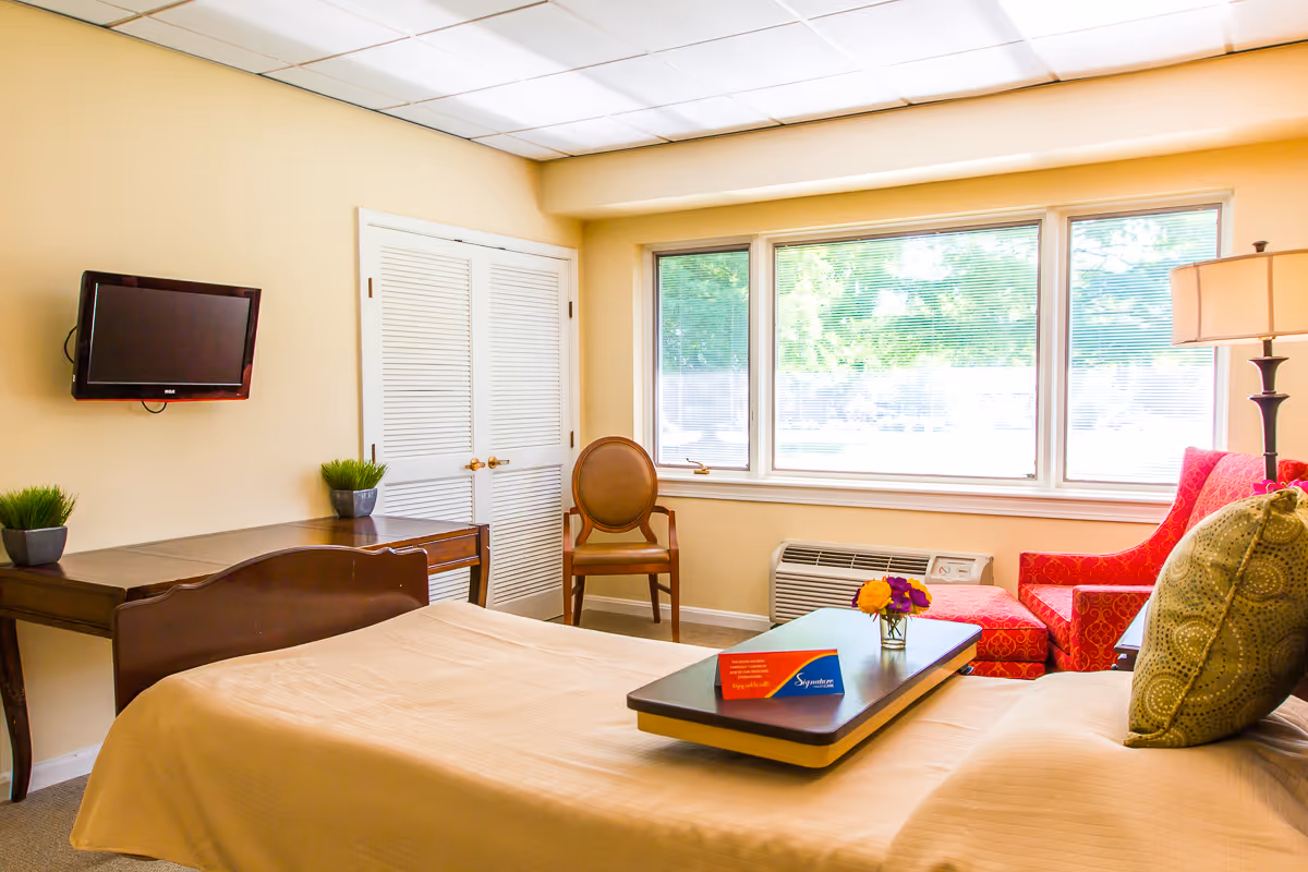 A bright and cozy bedroom in a senior living facility with a bed covered in beige bedding, a green patterned pillow, a wooden desk with two small potted plants, a wall-mounted flat-screen TV, a wooden chair, a red patterned armchair with a matching ottoman, a floor lamp, and large windows letting in natural light.