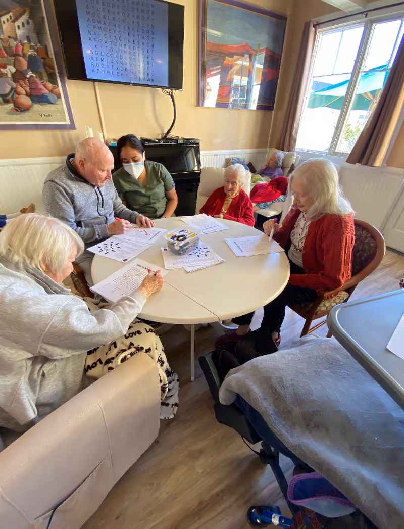 A group of elderly people and a caregiver wearing a mask sitting around a round table in a well-lit room. They are engaged in an activity involving papers and markers. A television screen on the wall displays a word search puzzle, and there are colorful paintings on the walls. Large windows let in natural light, and some elderly individuals are seated in armchairs near the window.