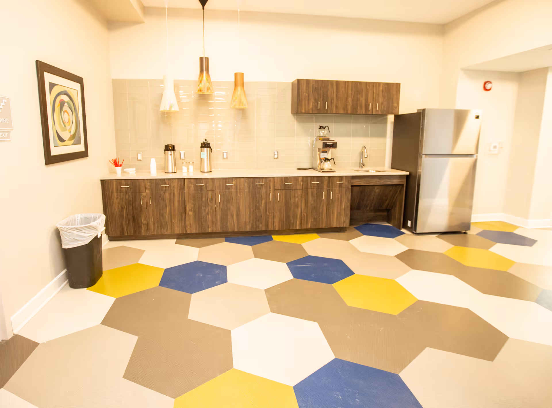 A kitchenette area with wooden cabinets, a stainless steel refrigerator, a coffee maker, and two coffee dispensers on the counter. The floor has a colorful hexagonal tile pattern in blue, yellow, white, and gray. There are three pendant lights hanging above the counter and a framed abstract artwork on the wall to the left. A trash can is placed near the wall under the artwork.